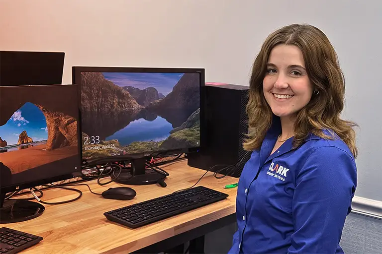 The CLARK Way | Clark Computer Services | CLARK Computer Services IT support specialist in blue company polo smiling at desk with dual-monitor workstation setup