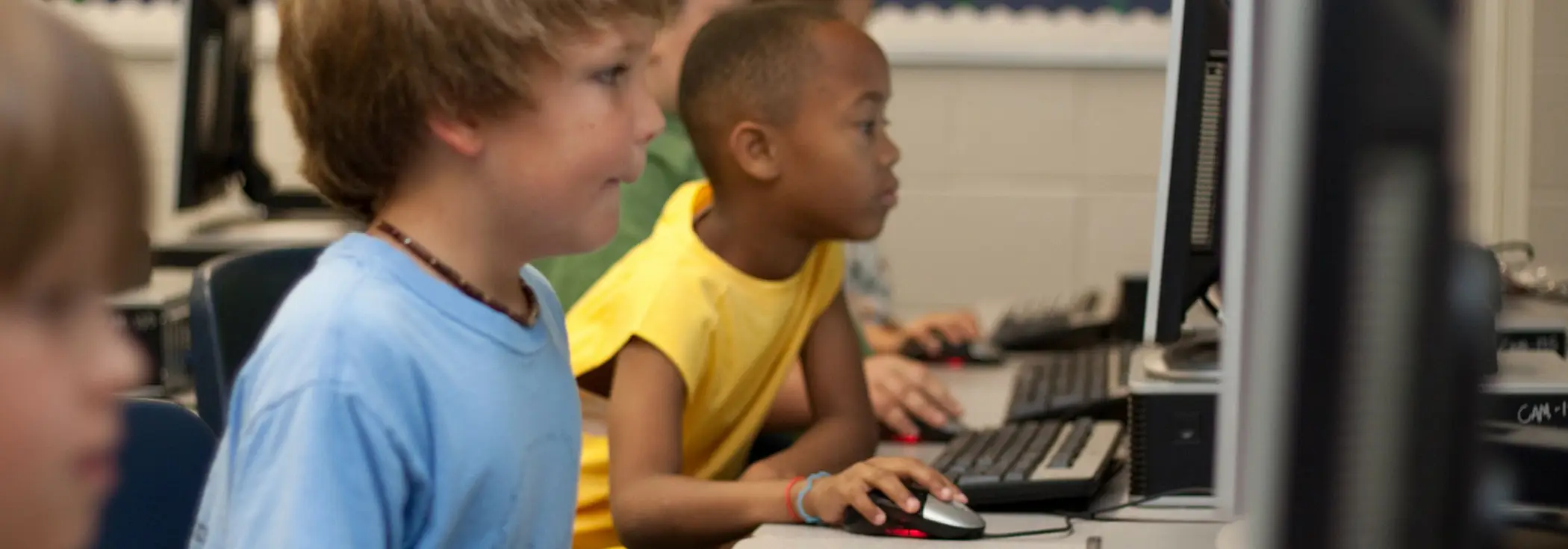 Two elementary school students, one in a light blue shirt and one in a yellow shirt, focused on using desktop computers in a community center