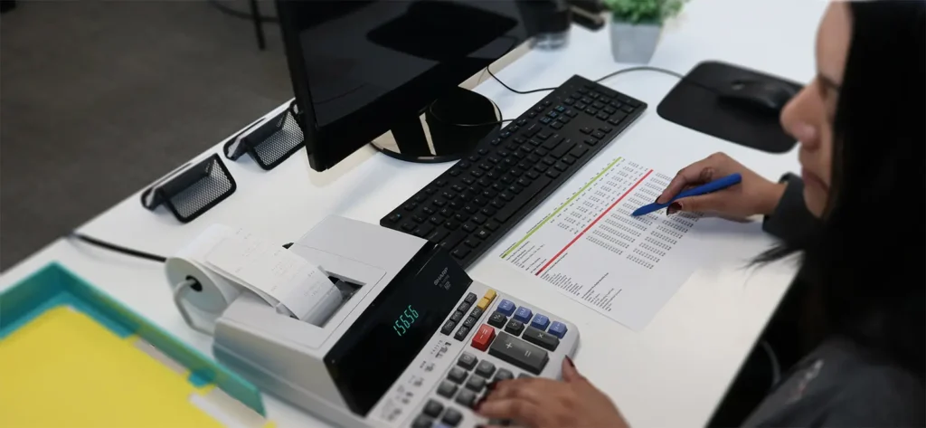 Finance IT Services | Secure, Reliable, & Compliant Support | Woman at a desk using an adding machine and reviewing a spreadsheet